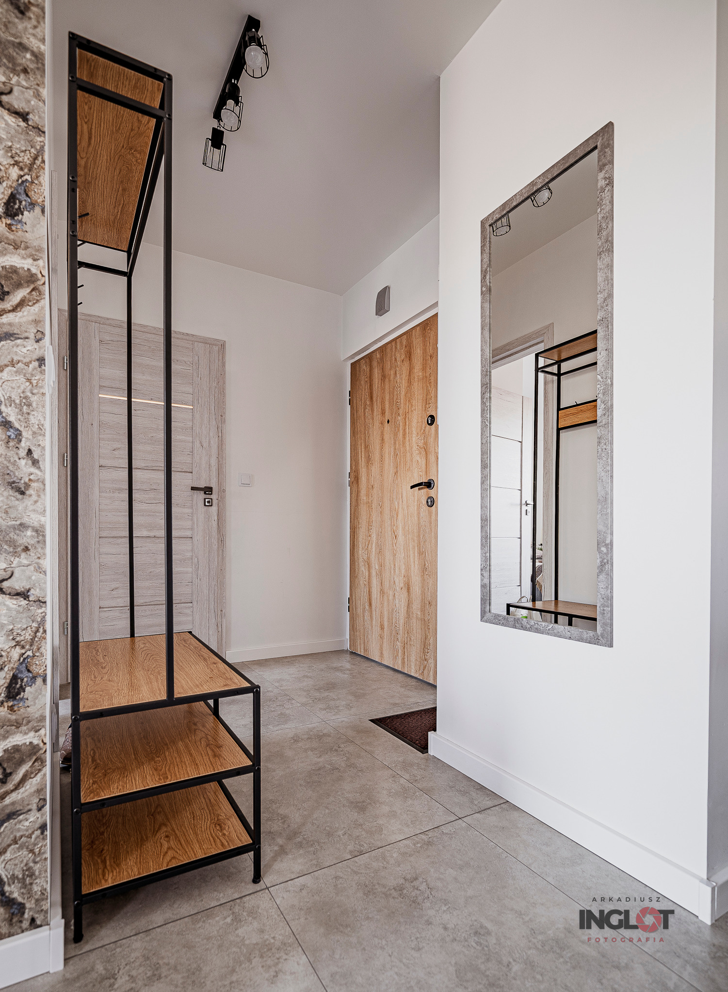 Nowoczesny przedpokój z szarą podłogą, drewnianymi drzwiami wejściowymi, lustrem w kamiennej ramie i metalowym regałem z drewnianymi półkami.