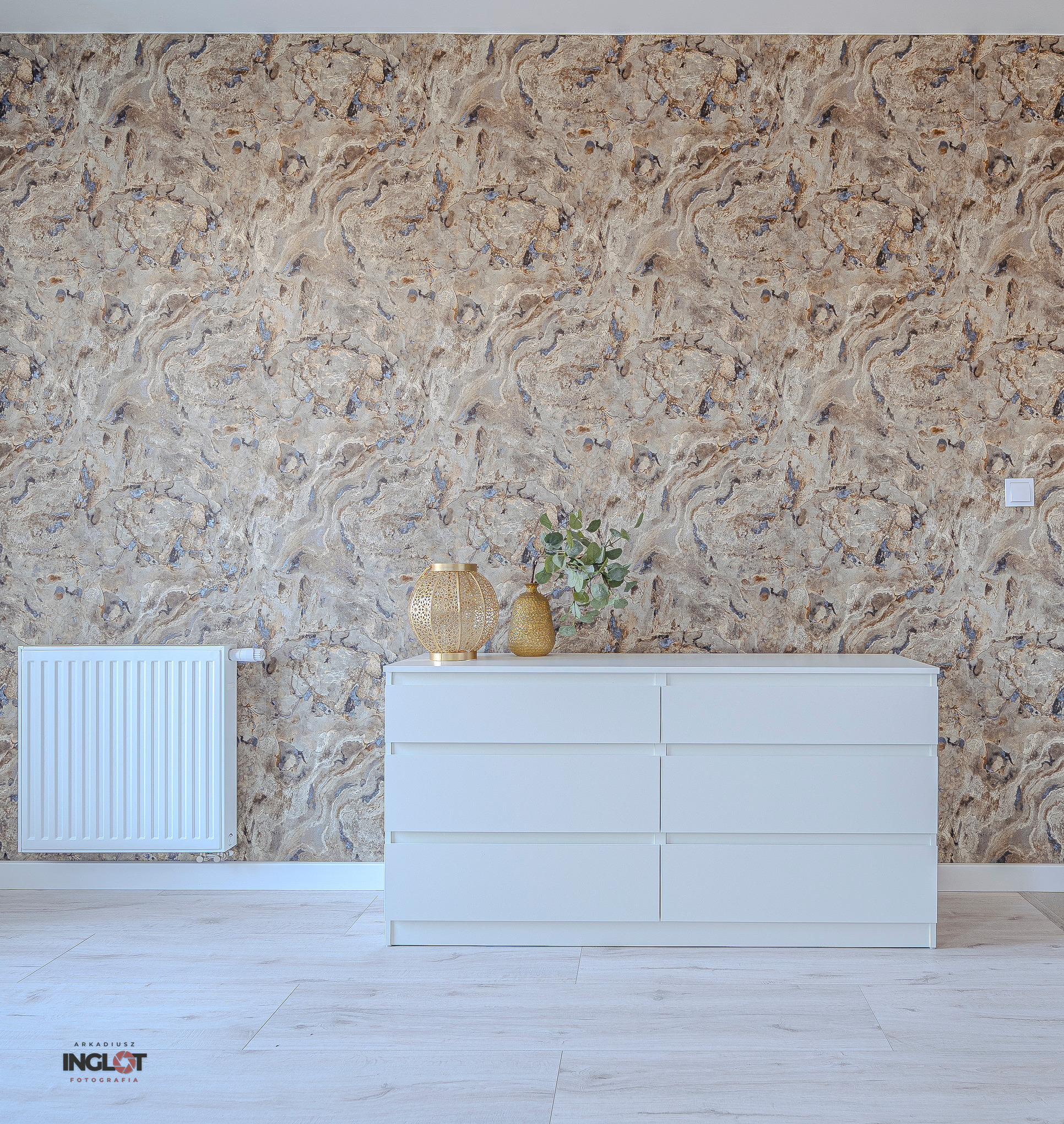 Jasna komoda z sześcioma szufladami, ozdobiona ażurową kulą i wazonem z zielonymi gałązkami na tle wzorzystej tapety, obok biały grzejnik.