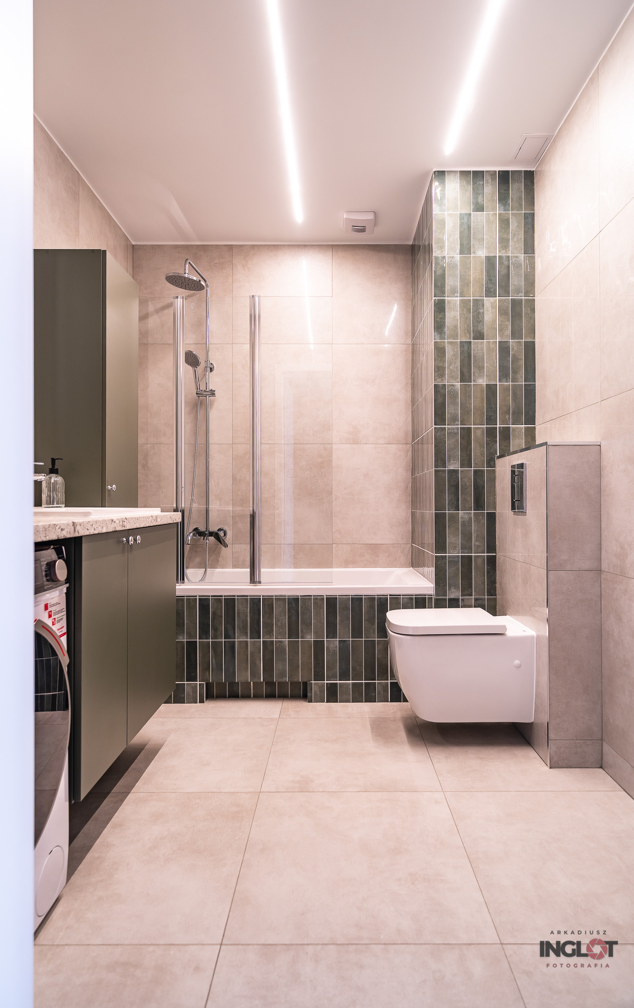 Nowoczesna łazienka z beżowymi płytkami podłogowymi i ściennymi, zielonymi szafkami, wanną z parawanem, wiszącą toaletą i podłużnymi lampami sufitowymi.