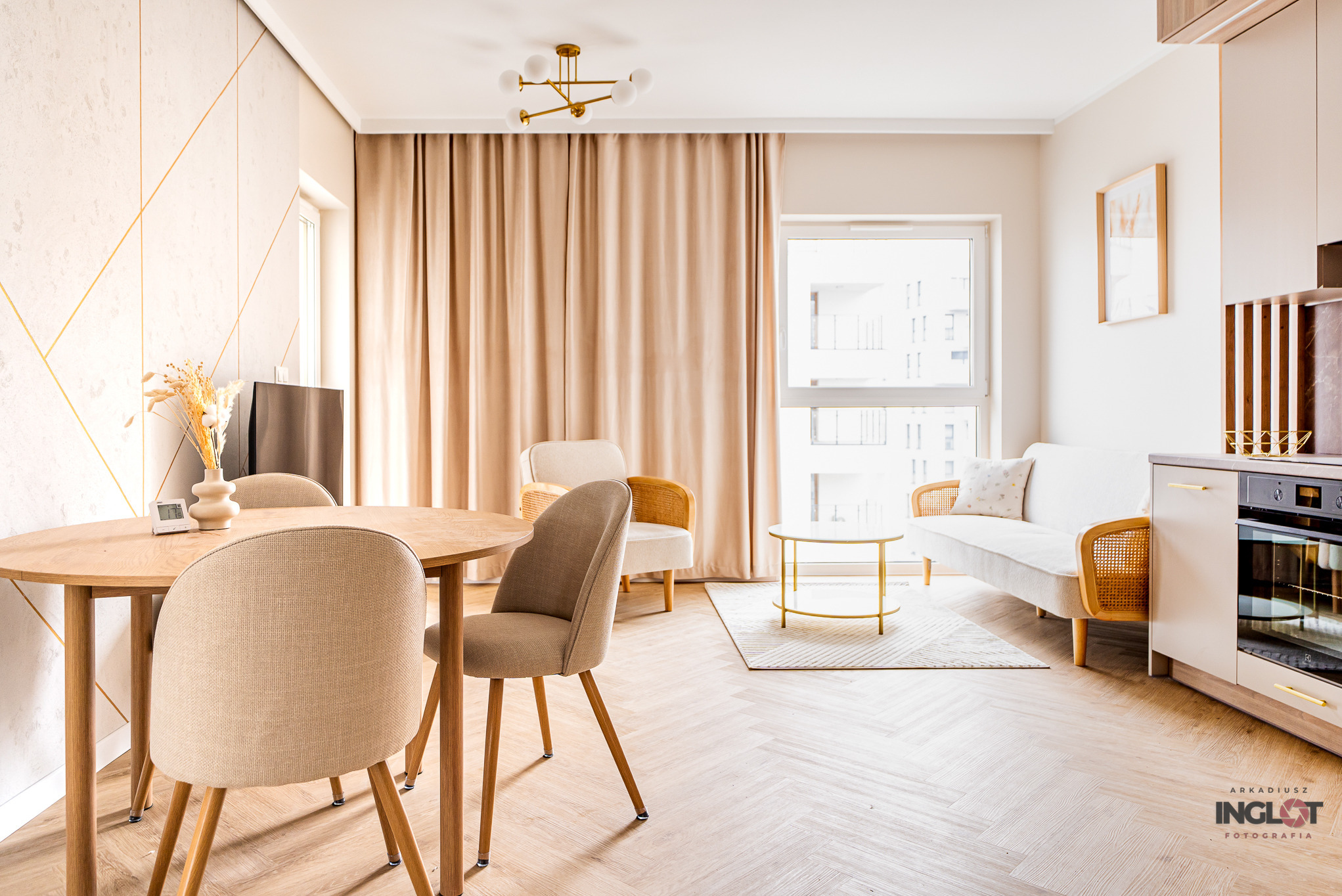 Jasne, nowocześnie urządzone wnętrze mieszkania z drewnianym stołem, krzesłami, kanapą z rattanowymi elementami, złotymi akcentami i parkietem ułożonym w jodełkę.