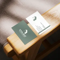 Dwie wizytówki na drewnianym podłokietniku krzesła. Jedna wizytówka w kolorze oliwkowym z napisem STOLARZ MEBLE NA WYMIAR i logo, druga biała z danymi kontaktowymi Lucjana Strancicha.