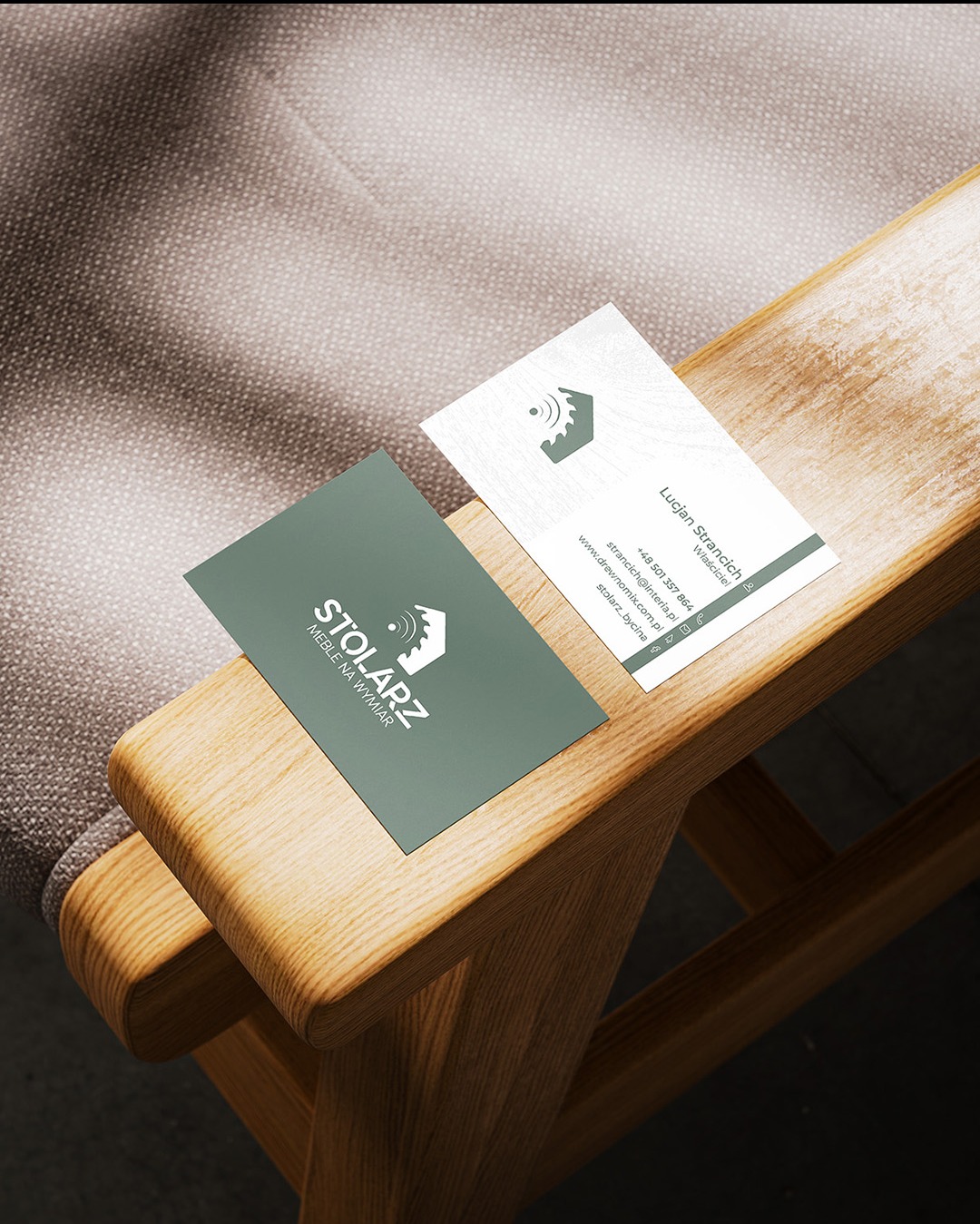 Dwie wizytówki na drewnianym podłokietniku krzesła. Jedna wizytówka w kolorze oliwkowym z napisem STOLARZ MEBLE NA WYMIAR i logo, druga biała z danymi kontaktowymi Lucjana Strancicha.
