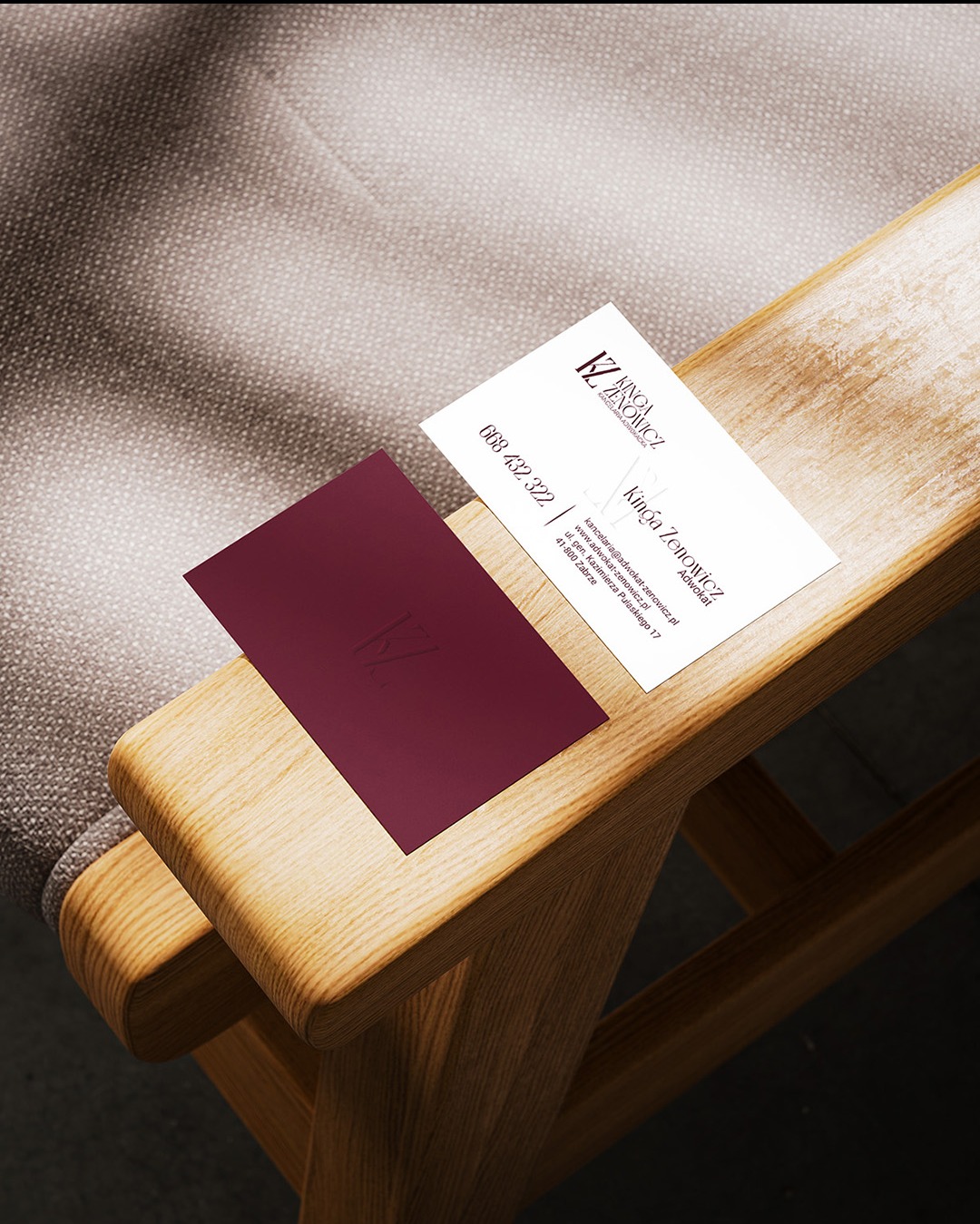 Dwie wizytówki, biała i bordowa z logo, leżą na drewnianym podłokietniku krzesła na tle szarej tapicerki.