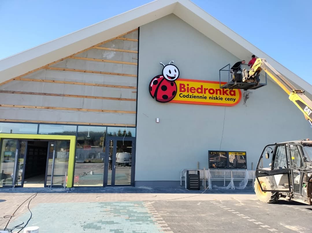Montaż logo Biedronki na elewacji budynku z użyciem podnośnika koszowego. Widoczne przygotowania do montażu i pracownicy w trakcie instalacji.