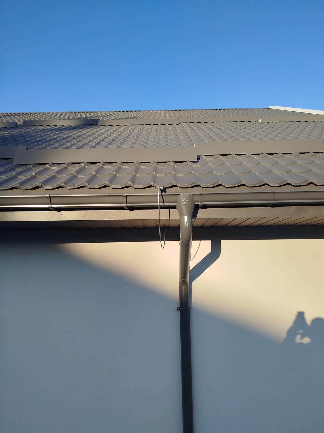 Szary system rynnowy zamontowany na budynku z blachodachówką w słoneczny dzień. Widoczny cień osoby fotografującej.