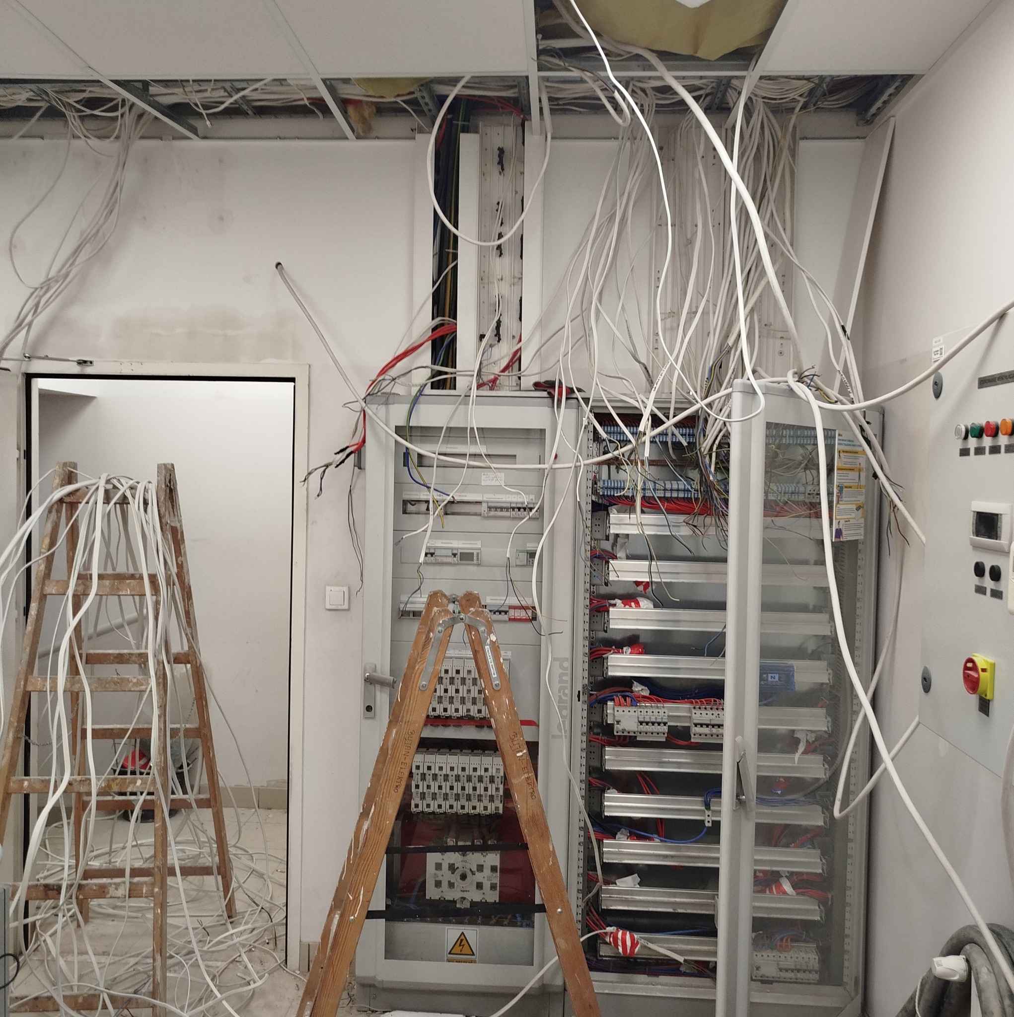 Rozbudowa rozdzielni elektrycznej z licznymi kablami zwisającymi z sufitu podwieszanego, drabiny malarskie w pomieszczeniu. Etap prac instalacyjnych.