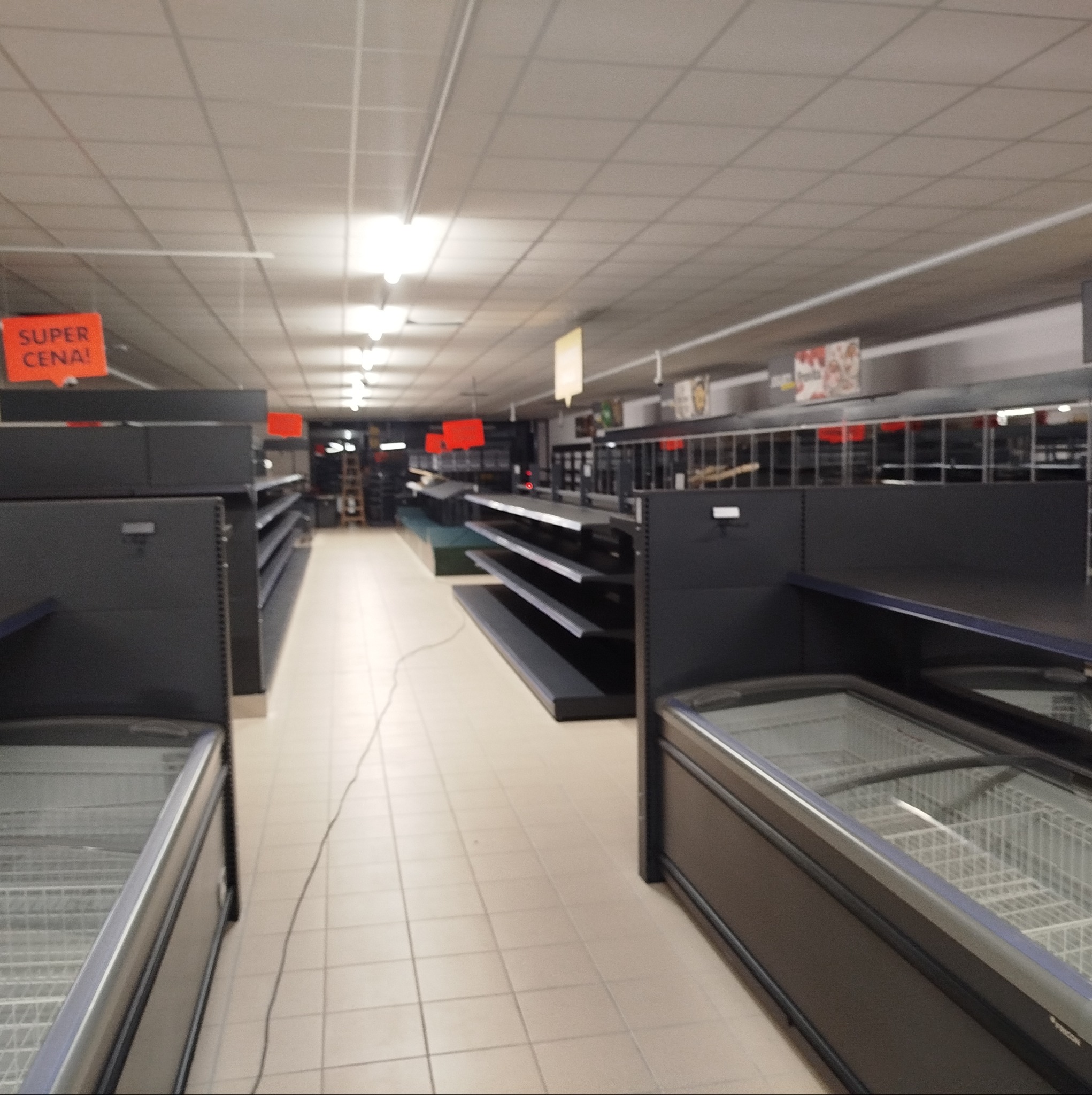 Oświetlenie sufitowe z rastrami w pustym sklepie, widoczne regały i chłodziarki. Pomarańczowe szyldy 'Super Cena!' podwieszone pod sufitem. Perspektywa wnętrza sklepu.