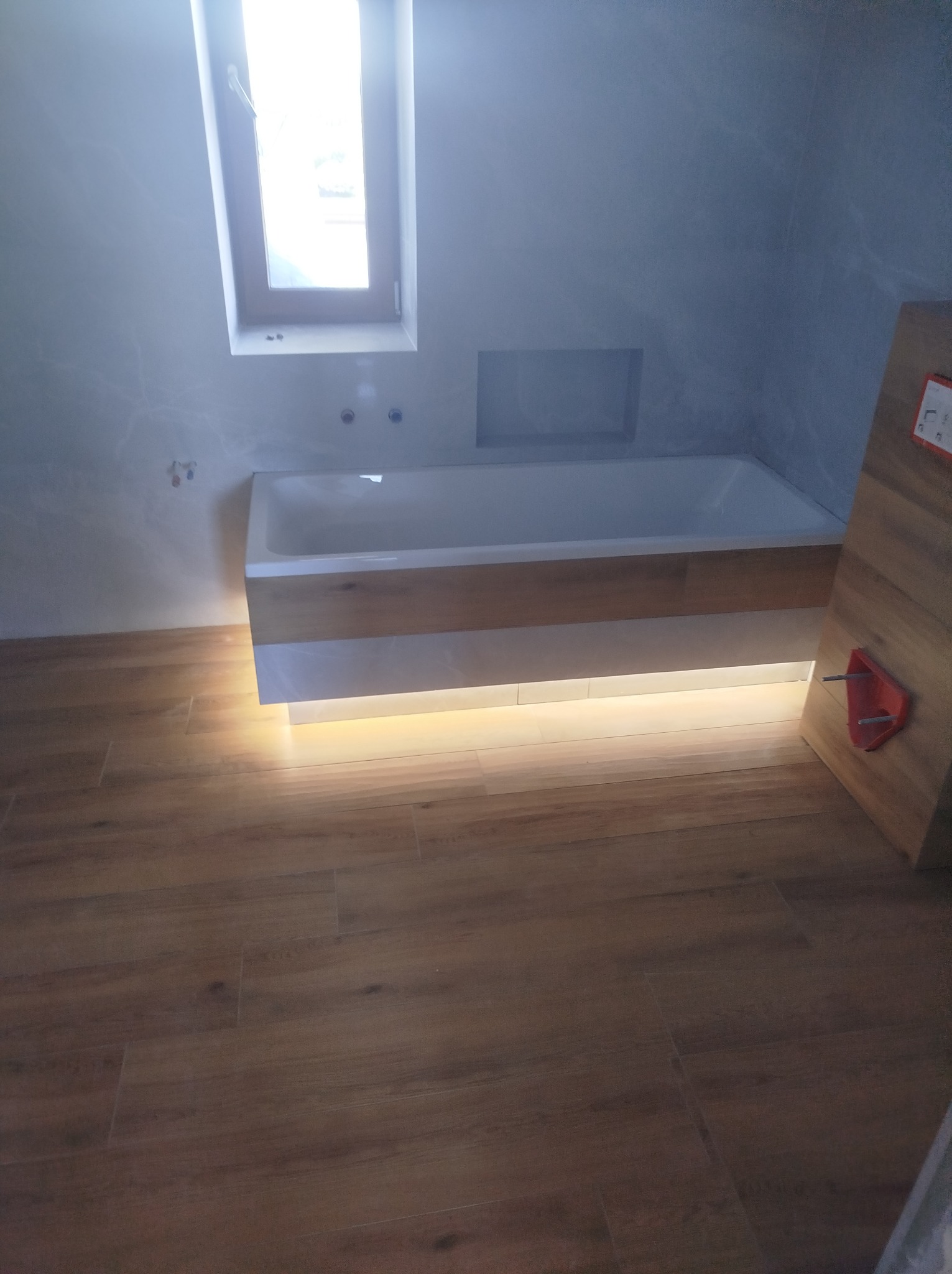 Nowoczesna wanna z podświetleniem LED w wykończonej łazience z drewnopodobną podłogą i oknem. Częściowo widoczny element montażowy po prawej stronie.