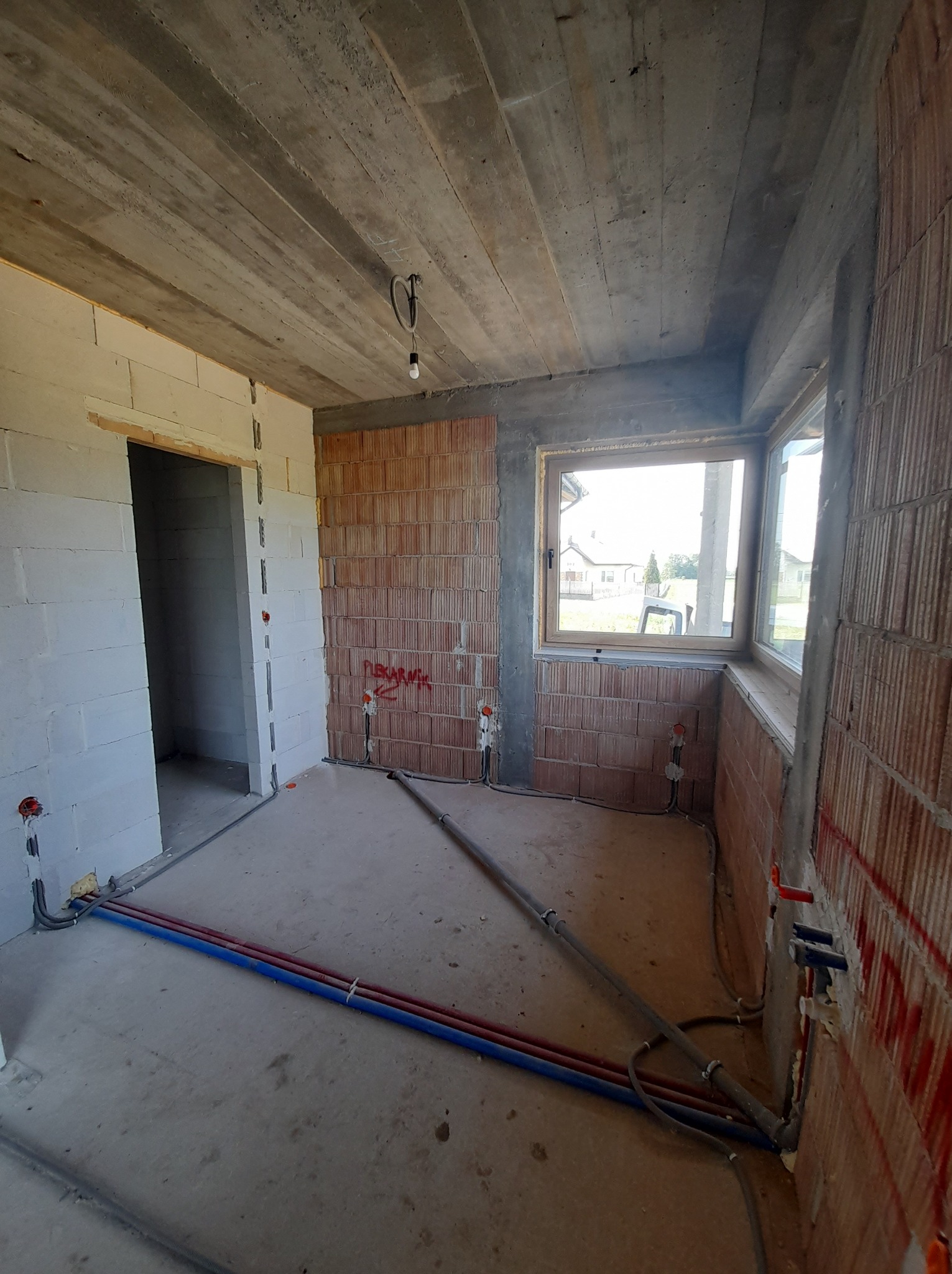 Surowy stan wnętrza z instalacjami: widoczne kable elektryczne w ścianach, rury na podłodze, okno z widokiem na zewnątrz. Ściany z cegły i betonu.