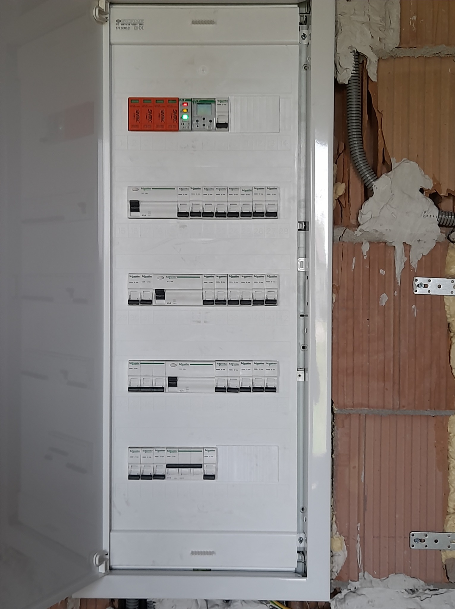 Otwarta, biała skrzynka z bezpiecznikami Schneider, zamontowana na ceglanej ścianie z widoczną instalacją elektryczną w peszlu. Widoczne zabezpieczenia przepięciowe.