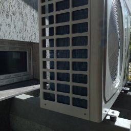 Eco-Free - Zewnętrzna jednostka klimatyzacji, widok z boku na betonowej podstawie, z widoczną kratką wentylacyjną i fragmentem elewacji budynku w tle.