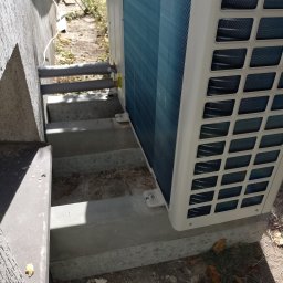 Eco-Free - Zewnętrzna jednostka klimatyzacji zamontowana na betonowych schodach przy ścianie budynku. Widoczne elementy instalacji, rury i mocowania, z bliska, z dołu.