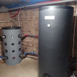 Eco-Free - Dwa zbiorniki akumulacyjne, szary i czarny, z podłączonymi rurami w piwnicy z ceglaną ścianą. Instalacja grzewcza, widoczne manometry i zawory.
