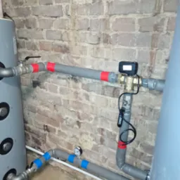 Szare, zaizolowane rury z zaworami i czujnikiem, połączone z szarym zbiornikiem, na tle ściany z cegły. Instalacja w piwnicy.