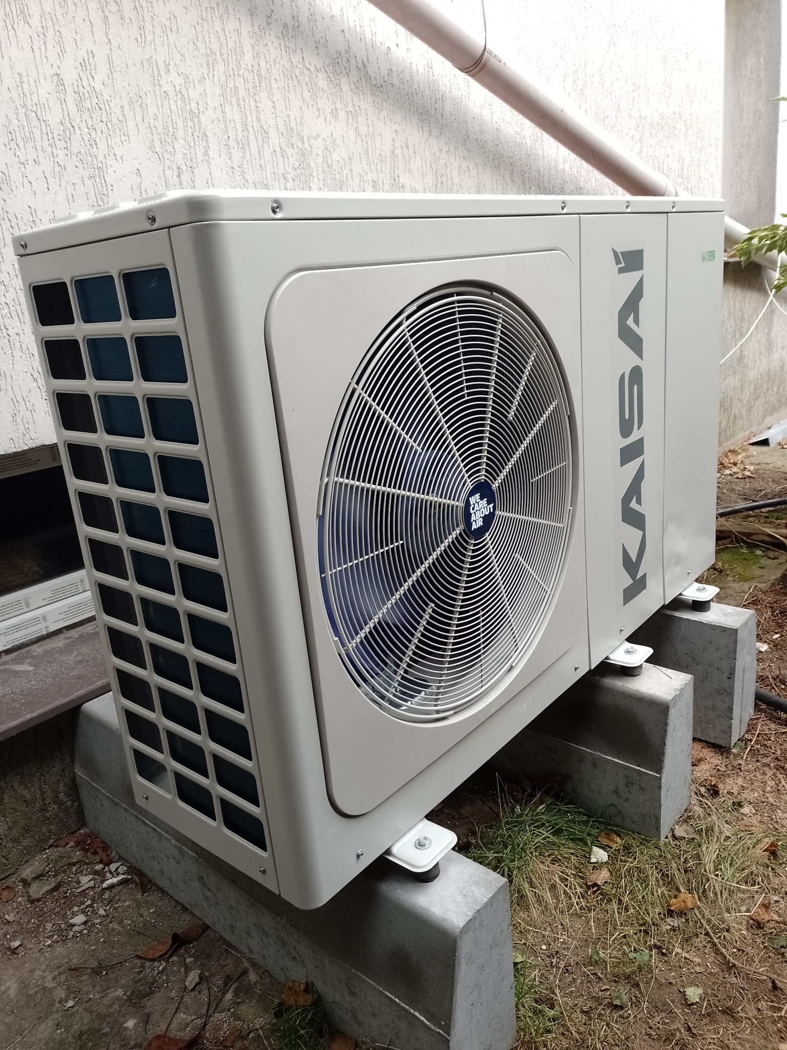 Zewnętrzna jednostka klimatyzacji Kaisai, umieszczona na betonowych wspornikach, stoi przy ścianie budynku. Widoczny wentylator i logo marki.