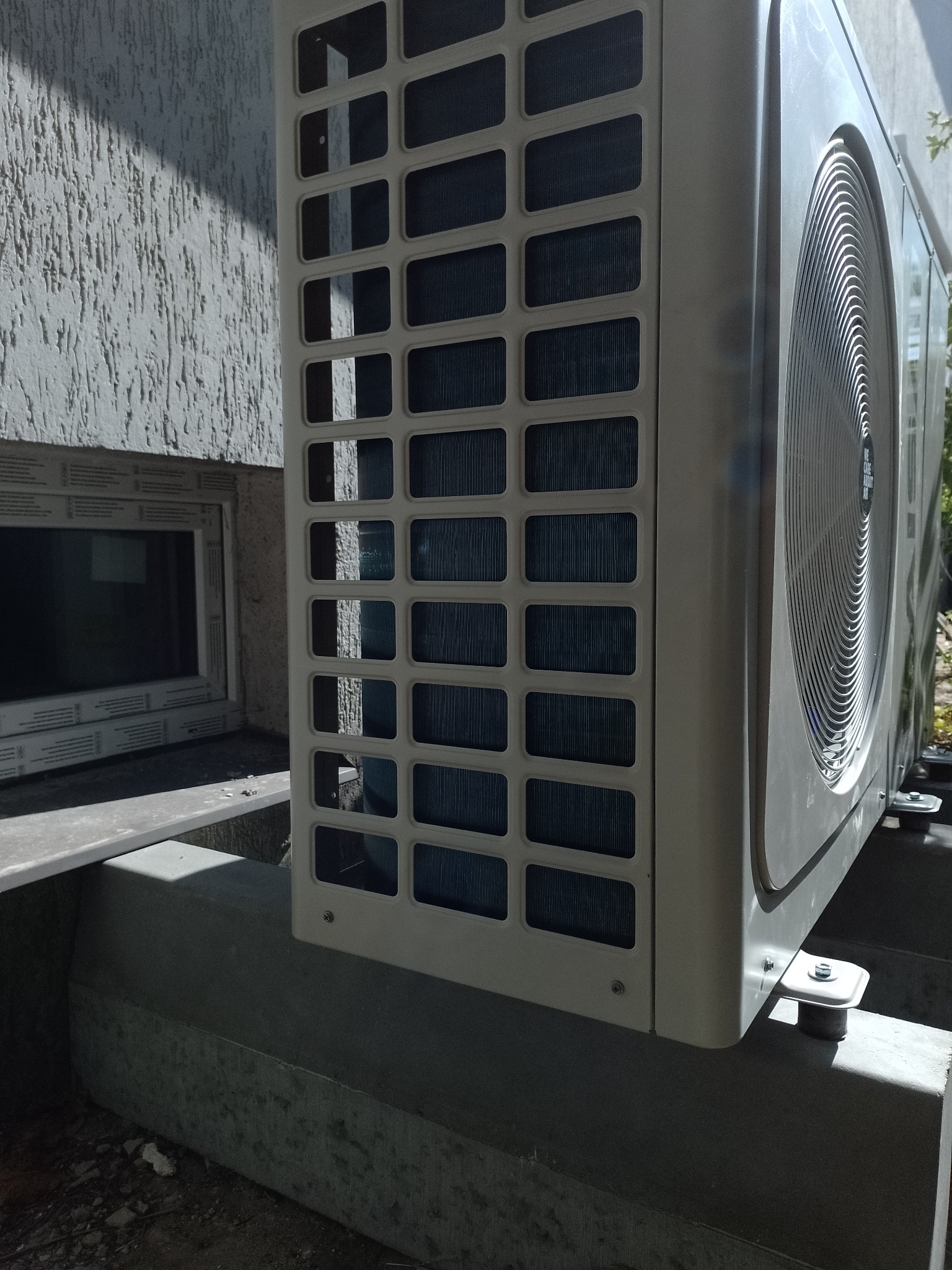 Zewnętrzna jednostka klimatyzacji, widok z boku na betonowej podstawie, z widoczną kratką wentylacyjną i fragmentem elewacji budynku w tle.