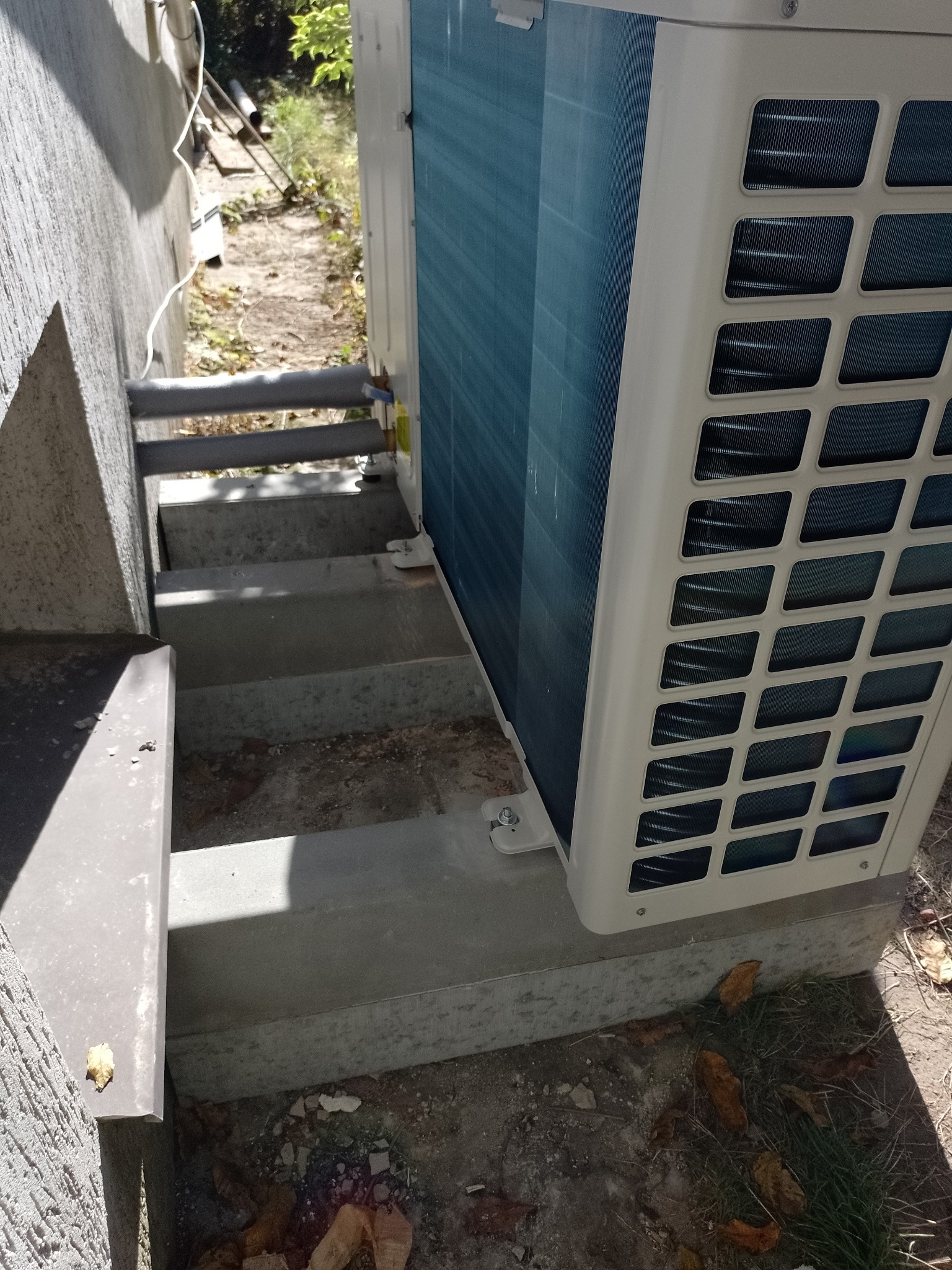 Zewnętrzna jednostka klimatyzacji zamontowana na betonowych schodach przy ścianie budynku. Widoczne elementy instalacji, rury i mocowania, z bliska, z dołu.