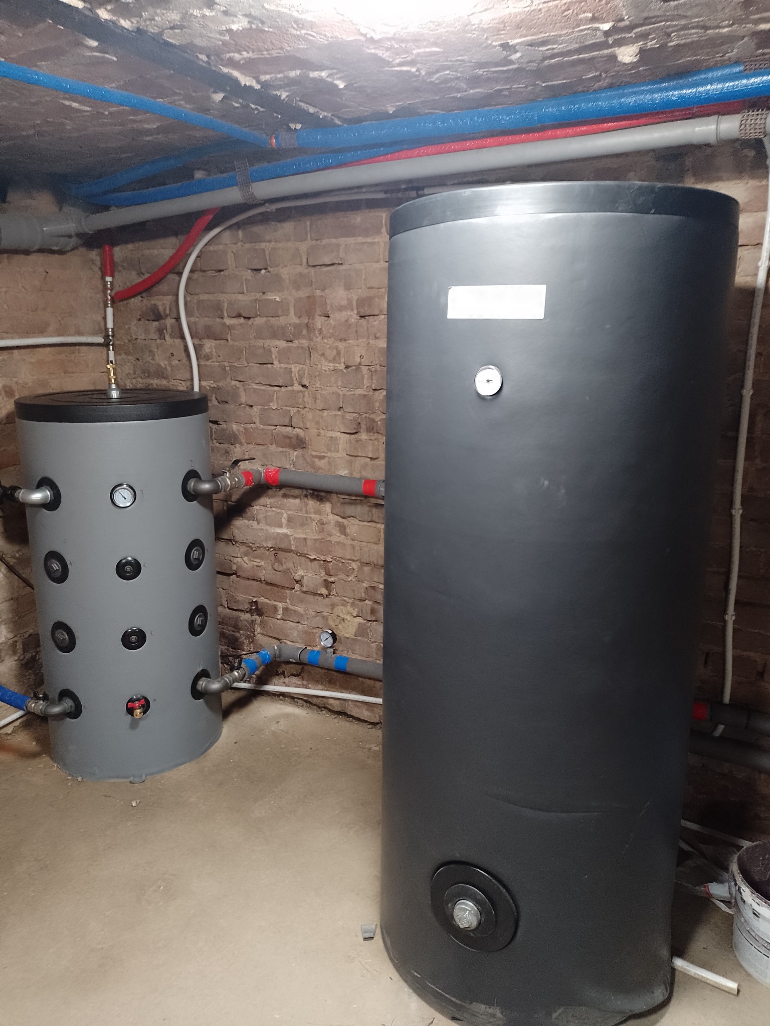 Dwa zbiorniki akumulacyjne, szary i czarny, z podłączonymi rurami w piwnicy z ceglaną ścianą. Instalacja grzewcza, widoczne manometry i zawory.