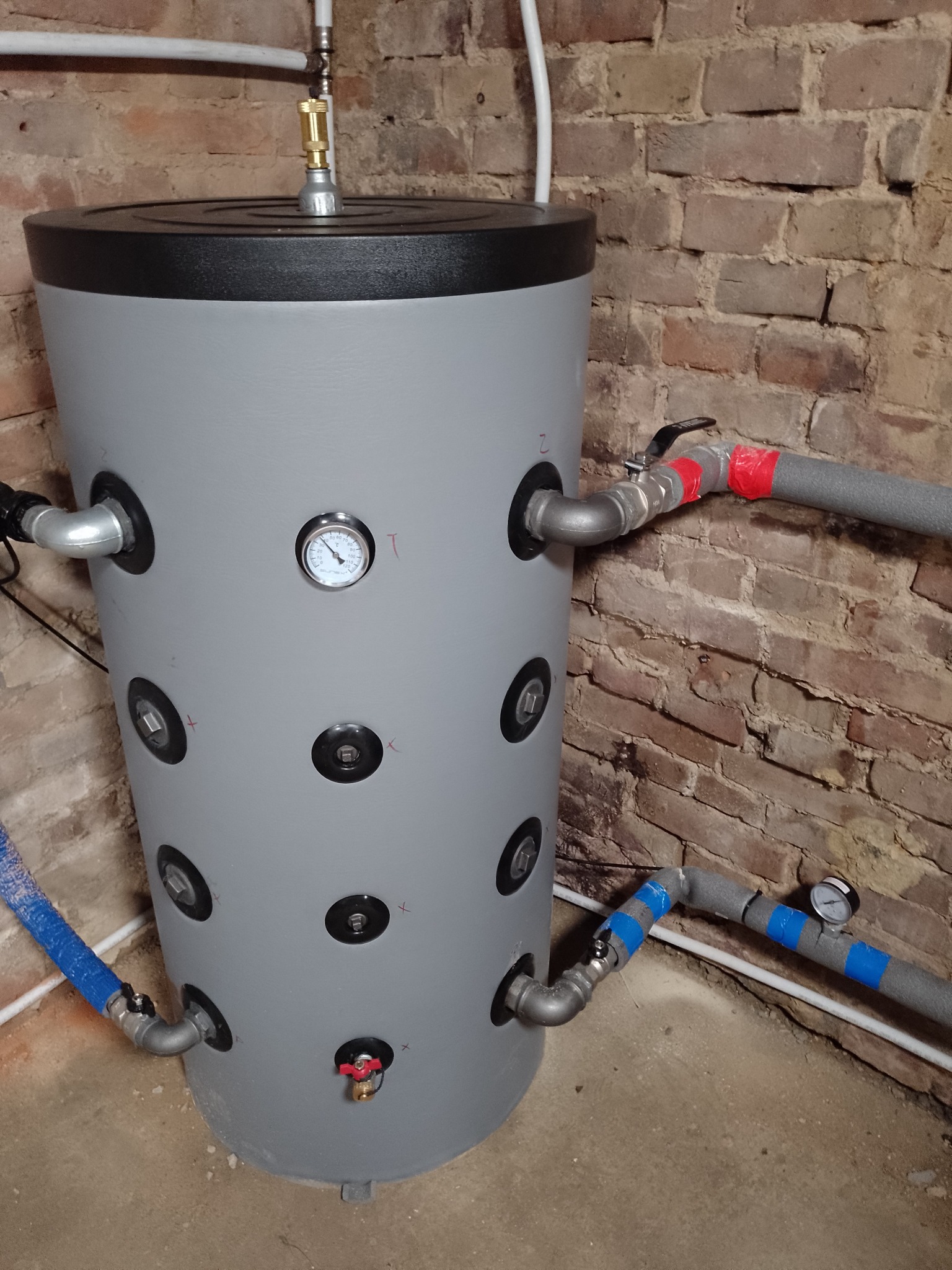 Szary zbiornik buforowy z licznymi przyłączami i manometrem, umieszczony w pomieszczeniu z ceglaną ścianą. Widoczne rury i izolacje.