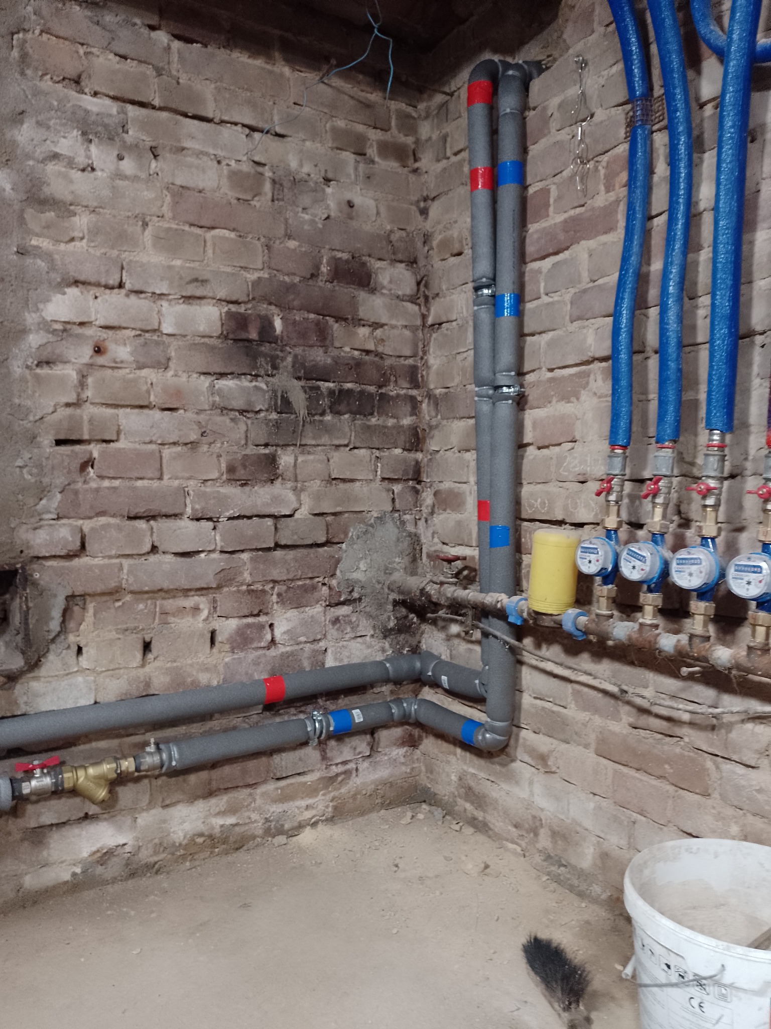 Instalacja wodna z izolowanymi rurami i licznikami na tle starej ceglanej ściany. Widoczny fragment podłogi i wiadro. Rury szare i niebieskie.