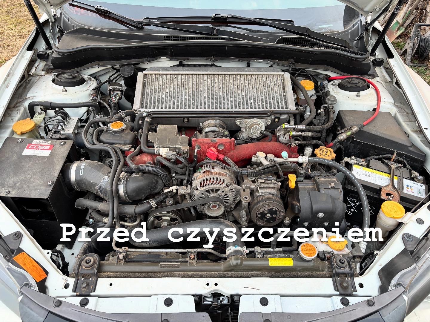 Brudny silnik samochodu Subaru przed czyszczeniem, widoczne zanieczyszczenia na elementach, w tym na intercoolerze i przewodach.
