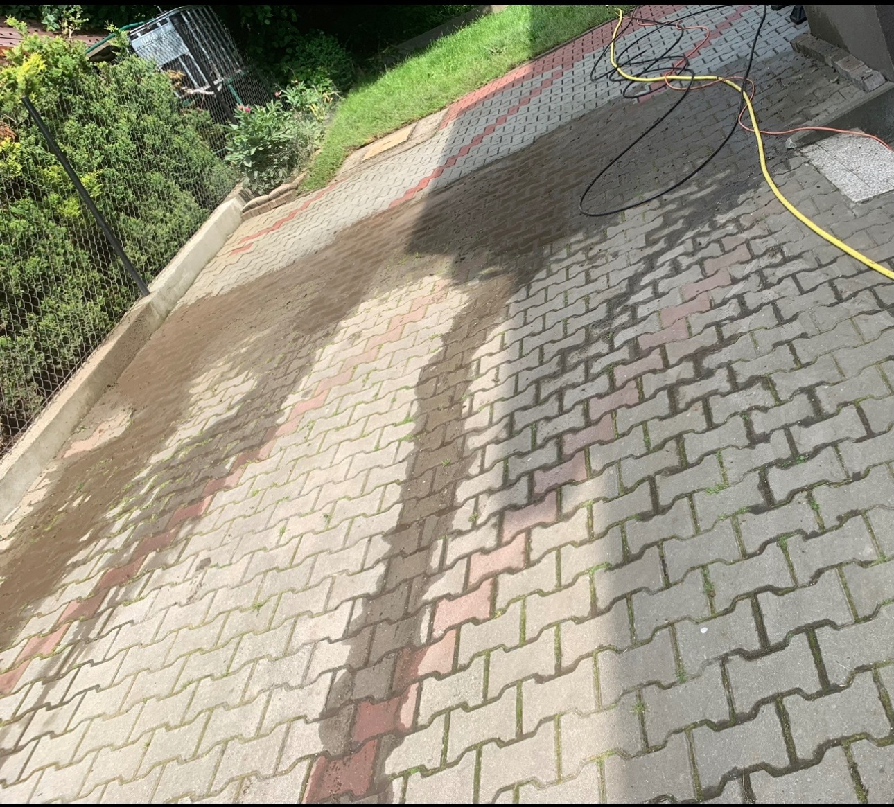 Częściowo umyta kostka brukowa w odcieniach szarości i czerwieni, widoczne mokre ślady po czyszczeniu, obok trawnik i krzewy, w tle zwinięte kable.