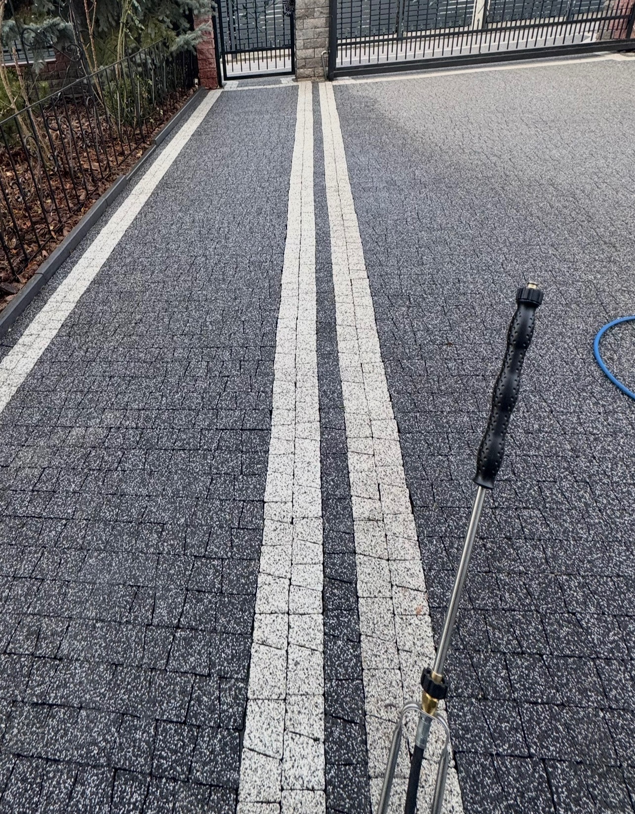 Czyszczenie kostki brukowej na podjeździe z użyciem myjki ciśnieniowej; widoczne dwie białe linie z kostki pośrodku szarej nawierzchni.