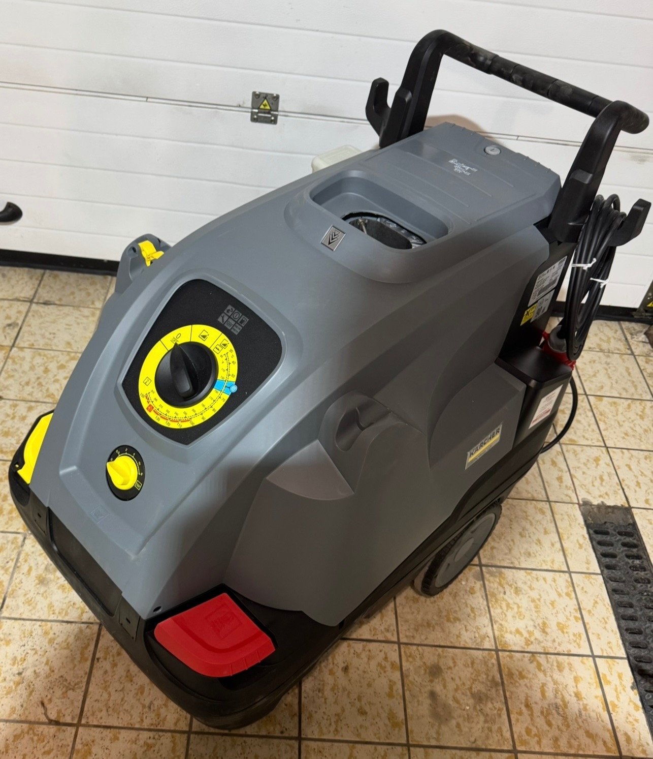 Profesjonalna myjka ciśnieniowa Karcher w kolorze szaro-czarnym z żółtymi akcentami, z widocznym panelem sterowania i uchwytem, stojąca na płytkach w pobliżu kratki odpływowej.