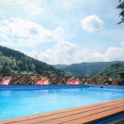 Ośrodek Wypoczynkowy Pod Dębem MARTA ŁUKAWSKA - Odkryty basen z błękitną wodą otoczony drewnianym tarasem i płotem z leżakami, w tle zielone wzgórza i błękitne niebo z chmurami.