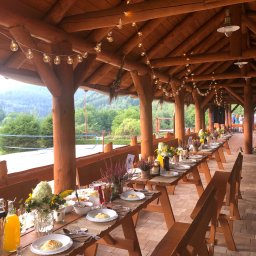 Ośrodek Wypoczynkowy Pod Dębem MARTA ŁUKAWSKA - Rustykalna wiata biesiadna z drewnianą konstrukcją i oświetleniem girlandami żarówkowymi, przygotowana do przyjęcia z długimi stołami udekorowanymi kwiatami, zastawą i przekąskami, w tle widok...