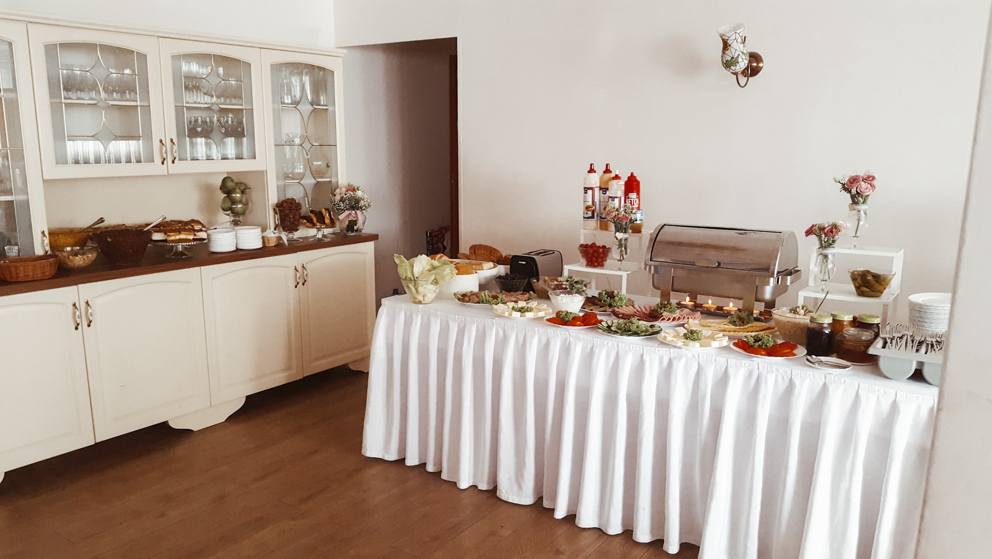 Bufet śniadaniowy z różnorodnymi potrawami, wędlinami, serami, warzywami, pieczywem, słodkościami i napojami, ustawiony na stole z białym obrusem, w tle kredens z szklankami i dekoracjami.