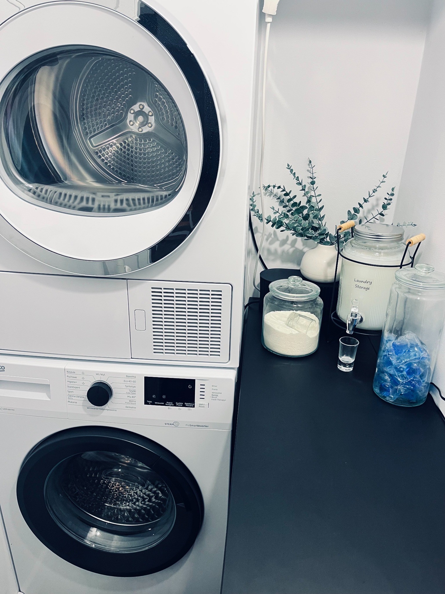 Stos pralki i suszarki w jasnym pomieszczeniu, obok dekoracyjne słoje z detergentami i rośliną, minimalistyczny design.