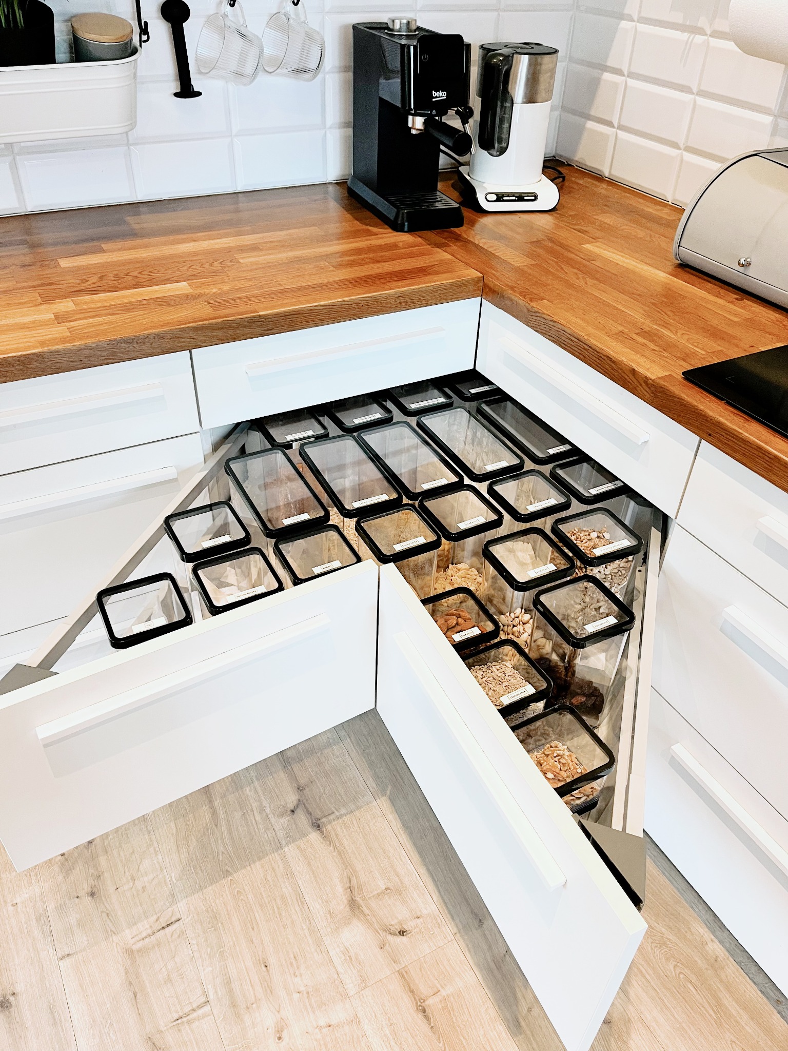 Wnętrze szafki narożnej w kuchni z wysuwanymi półkami, wypełnionymi przezroczystymi pojemnikami z różnymi rodzajami żywności, takimi jak orzechy, ziarna i suszone owoce, z drewnianym blatem...