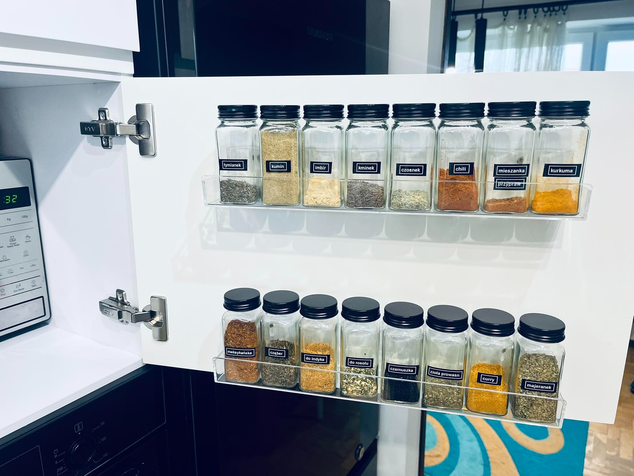Szafka kuchenna z otwartymi drzwiczkami, na których zamontowane są dwie półki z przezroczystego tworzywa, wypełnione szklanymi słoiczkami z czarnymi zakrętkami i etykietami, zawierającymi przyprawy...