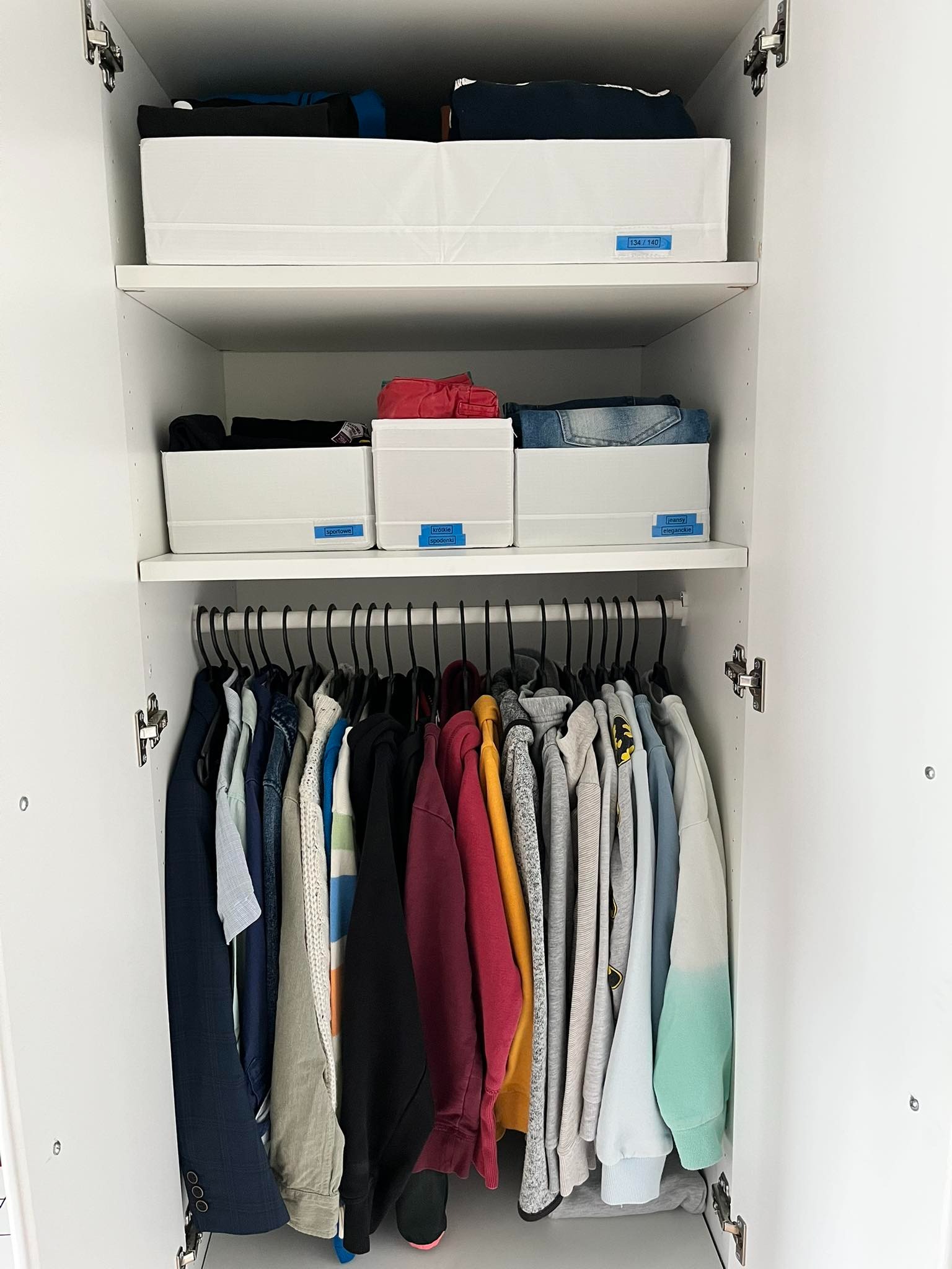 Uporządkowana szafa z ubraniami posegregowanymi w białych pojemnikach z etykietami i wiszącymi na czarnych wieszakach.