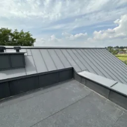 Szary dach z blachy na domu w Mietniowie, widoczne obróbki blacharskie i komin wentylacyjny na tle błękitnego nieba.