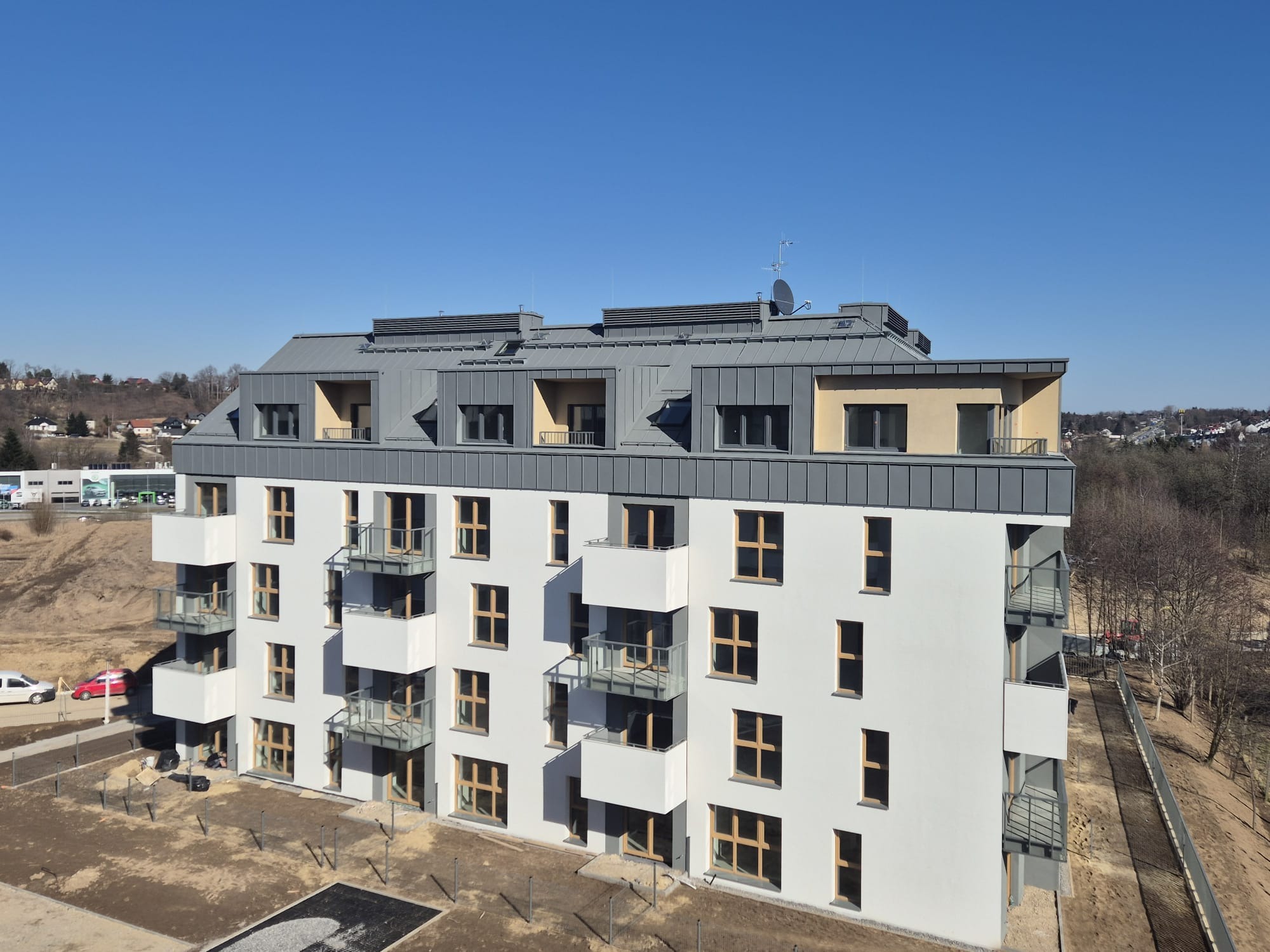 Nowoczesny budynek wielorodzinny z szarą blachą na dachu, balkonami z przezroczystymi balustradami i białymi elewacjami, widziany z lotu ptaka w słoneczny dzień.