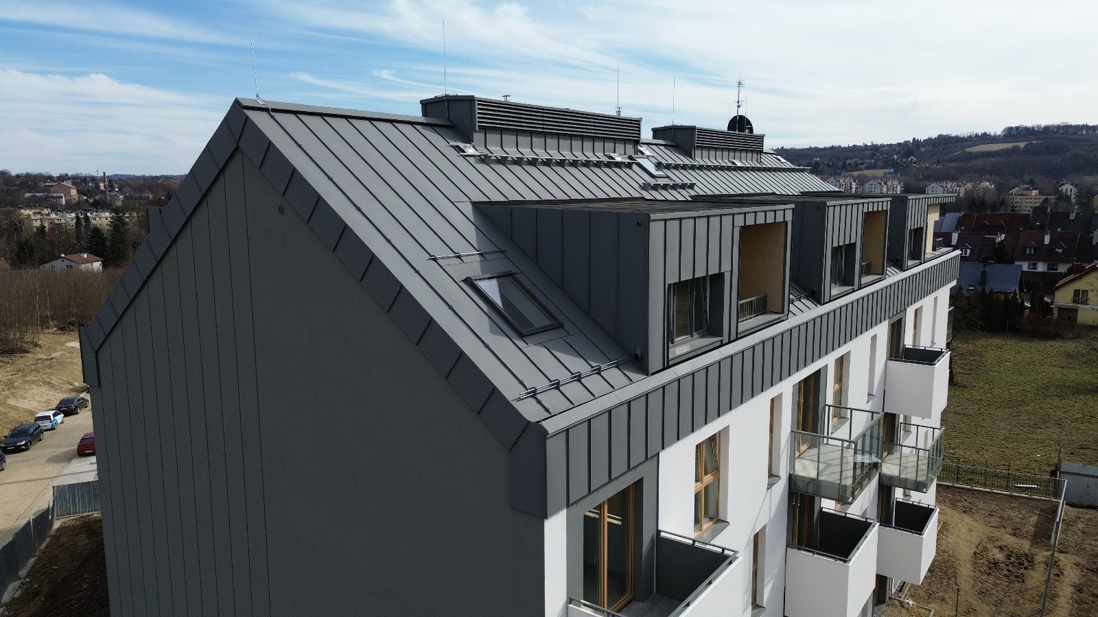 Nowoczesny budynek wielorodzinny z szarym dachem z blachy na rąbek stojący, lukarnami i oknami dachowymi, widok z góry na tle zabudowy miejskiej.