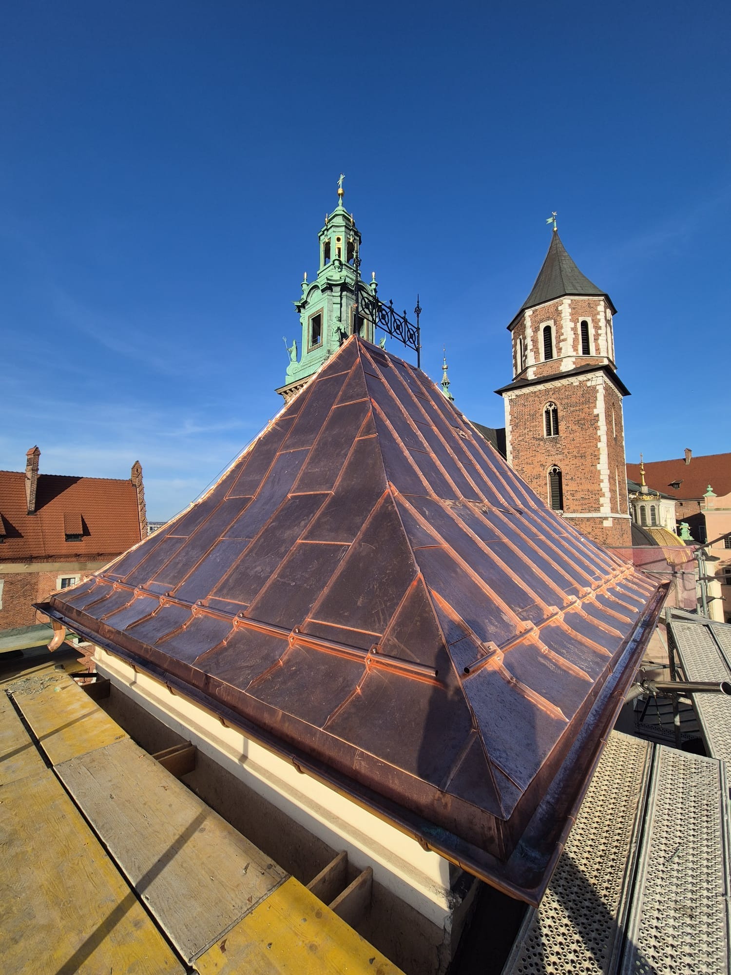 Miedziany dach piramidalny na tle zabytkowych wież w Krakowie, widok z rusztowania podczas prac.