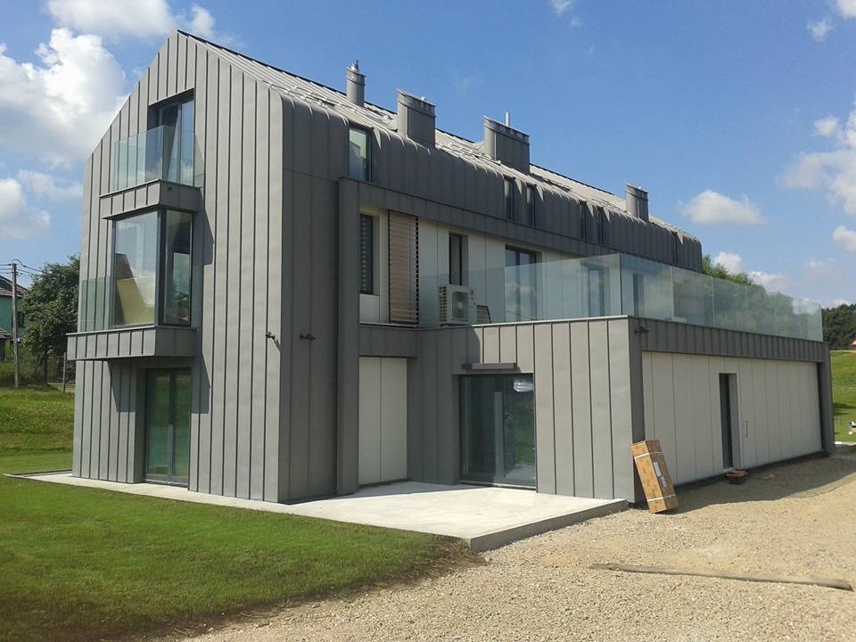 Nowoczesny dom jednorodzinny z szarą blachą na elewacji i dachu, balkony z szklanymi balustradami, widok z zewnątrz na trawnik i podjazd.