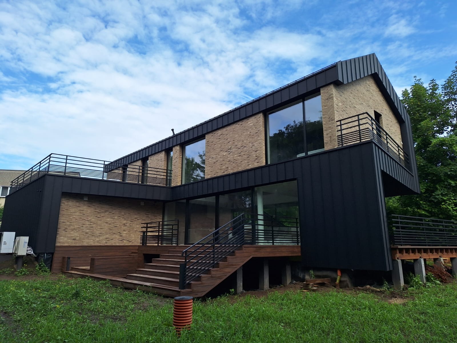 Nowoczesny dom z płaskim dachem wykończonym blachą w kolorze antracytowym, elewacja z cegły klinkierowej, duże okna, balustrady balkonowe z czarnych prętów, drewniany taras i schody wejściowe.