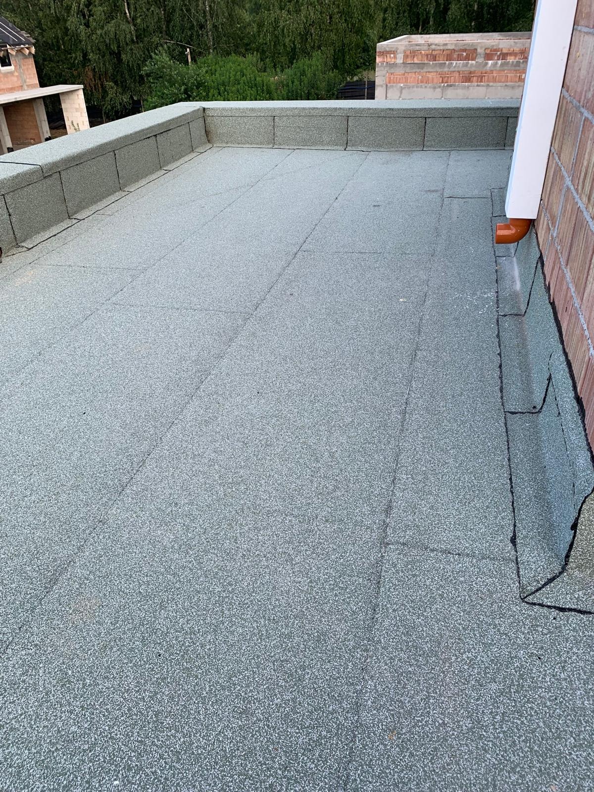 Wykończony płaski dach pokryty papą z widoczną attyką i fragmentem rynny spustowej przy ceglanej ścianie budynku w tle zielone drzewa.