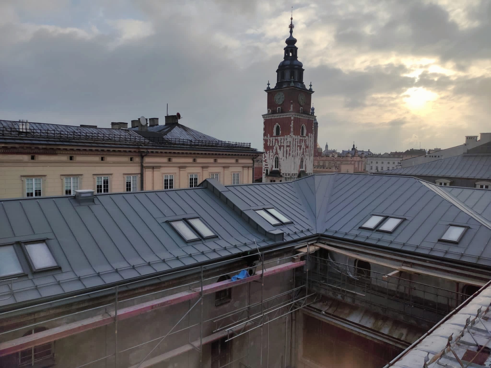 Nowy, szary dach z oknami połaciowymi na tle zabytkowej wieży zegarowej w Krakowie, widoczne rusztowania.