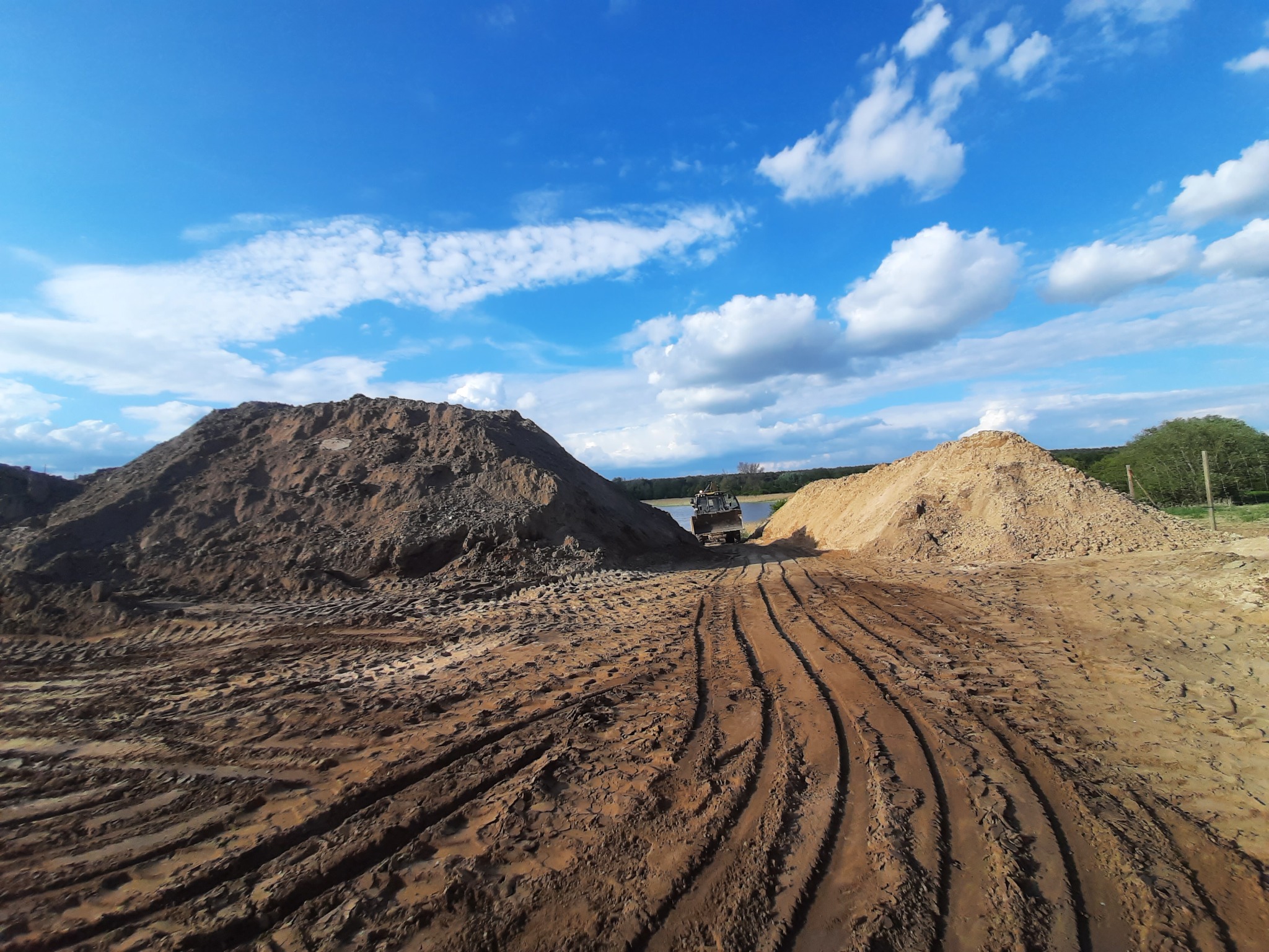 Koparka pomiędzy dwoma pryzmami ziemi na placu budowy, widoczne ślady opon na ubitej ziemi, błękitne niebo z białymi chmurami w tle.