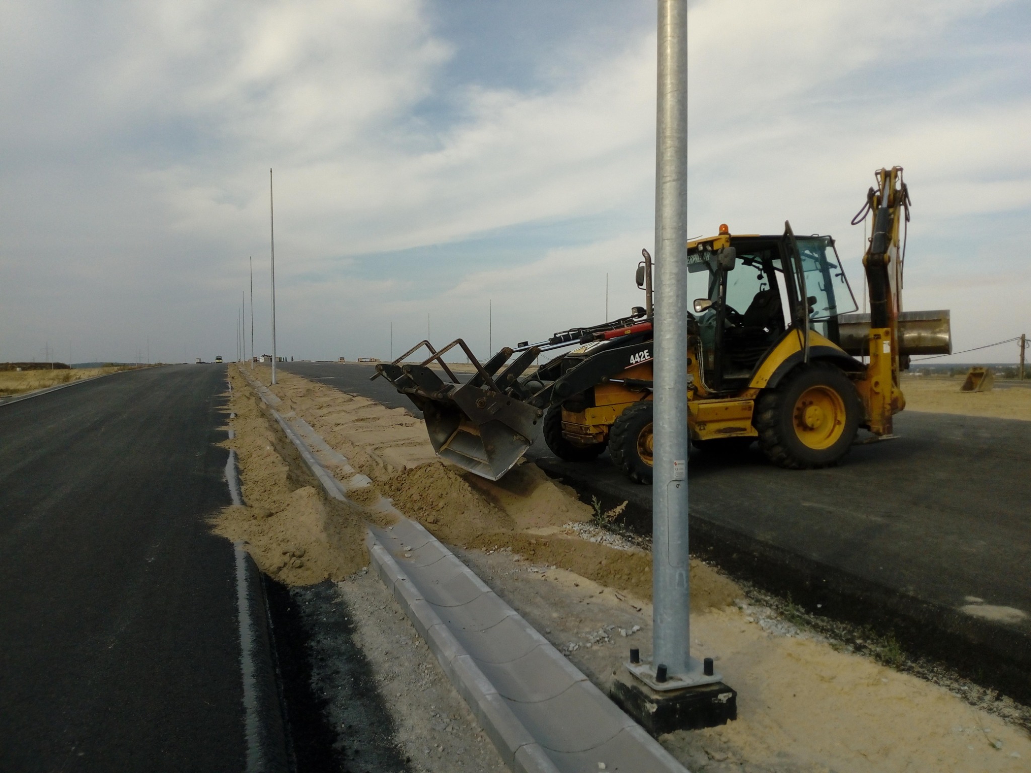 Żółta koparko-ładowarka Caterpillar 442E wyrównuje pobocze nowej drogi S5, widoczne krawężniki i słupy oświetleniowe.