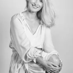 Portret uśmiechniętej kobiety w jasnej sukience, w czerni i bieli, siedzącej z rękami na kolanie, z widocznym pierścionkiem na palcu.