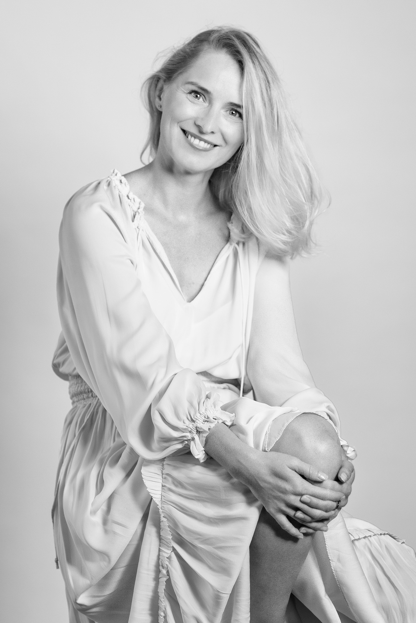 Portret uśmiechniętej kobiety w jasnej sukience, w czerni i bieli, siedzącej z rękami na kolanie, z widocznym pierścionkiem na palcu.