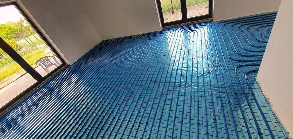 Niebieskie rury ogrzewania podłogowego ułożone na folii izolacyjnej z siatką, widok z góry na dużą powierzchnię podłogi w nowym domu, z oknami w tle.