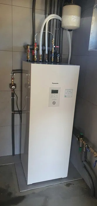 Biała pompa ciepła Panasonic zamontowana w pomieszczeniu technicznym, widoczne rury doprowadzające i odprowadzające wodę, manometr na rurze z lewej strony.