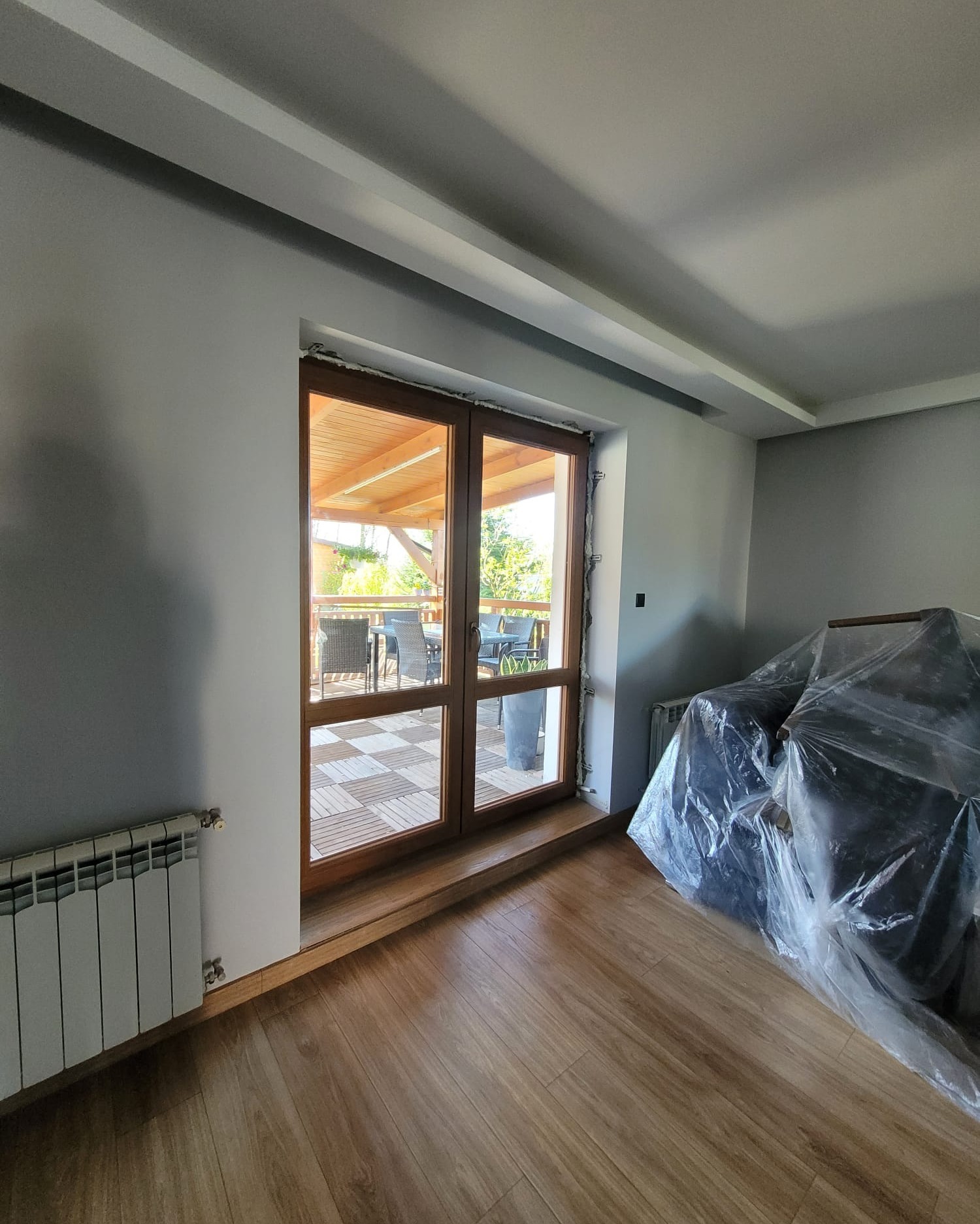 Wnętrze pokoju z drewnianą podłogą i grzejnikiem, widok na taras przez dwuskrzydłowe, drewniane drzwi balkonowe. Na zewnątrz drewniany taras z meblami ogrodowymi.